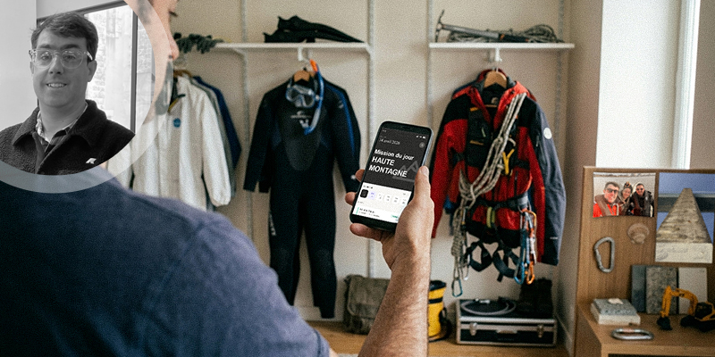 Person holding a smartphone in a climbing gear shop, with jackets, harnesses, and gear in the background.
